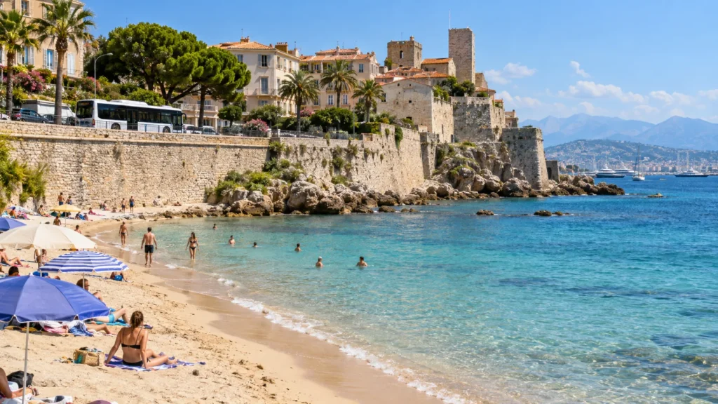 Sandy beach below stone ramparts, turquoise water and a bus on the seafront road, Antibes